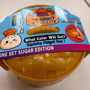 Puede incluir: Un recipiente redondo de plástico amarillo con textura tejida, que presenta una funda de cartón azul etiquetada como Squishy Bun Rainbow Mystery. El empaque incluye textos como What Color Will Get? Squishy Fidget Fun! y One Set Sugar Edition.
