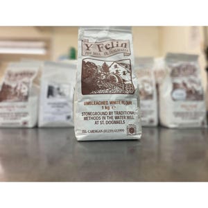 May include: Several white paper bags of flour are arranged on a surface. The central bag displays a brown illustration of a water mill and text: "Unbleached White Flour 1 kg e". It also states "Stoneground by Traditional Methods in the Water Mill at St. Dogmaels".