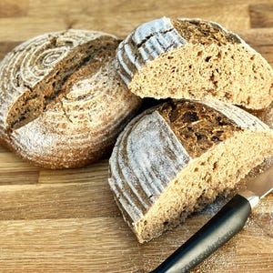 May include: Three loaves of freshly baked bread, one whole and two sliced, sit on a wooden surface. The bread has a dark brown crust with a spiral design on top. Sliced loaves reveal a light brown interior. A black-handled knife rests nearby.