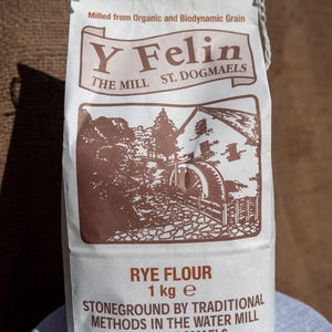 May include: A white paper bag of rye flour, featuring brown text and a detailed illustration of a water mill. The bag is labelled "Y Felin" and "Rye Flour 1 kg e". The text also includes "Stoneground by Traditional Methods in the Water Mill at St. Dogmaels."