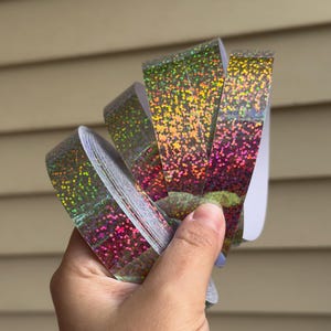 Puede incluir: Una mano sostiene una pila de rollos de cinta iridiscente y brillante. La cinta hace la transición del verde al rosa y al dorado, con un patrón moteado. El fondo es una superficie clara, con listones horizontales.