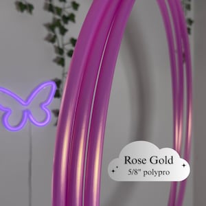 Puede incluir: Hula hoop de oro rosa hecho de tubo de polipropileno de 1,6 cm. El aro es de color rosa vibrante y tiene un acabado liso y brillante. Una mariposa de neón púrpura está en el fondo.