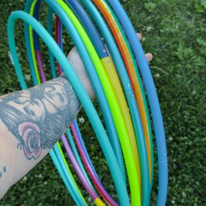 Peut inclure: Une collection de plusieurs cerceaux circulaires de différentes couleurs vives, notamment bleu sarcelle, jaune fluo, orange et bleu. Chaque cerceau est fabriqué à partir de tubes en plastique segmentés reliés par des rivets métalliques.