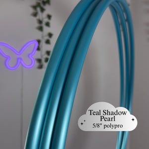 Puede incluir: Primer plano de un hula hoop de color turquesa. El texto en la imagen dice "Teal Shadow Pearl 5/8" polypro". El aro es de un vibrante tono turquesa, con un acabado suave y metálico. El fondo incluye una mariposa de neón púrpura y vegetación.