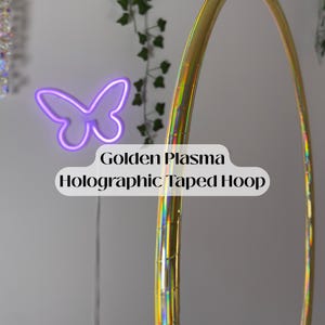 May include: A golden holographic taped hoop is featured in this image. The hoop is curved and has a shiny, reflective surface. The text "Golden Plasma Holographic Taped Hoop" is displayed. A purple neon butterfly is in the background.
