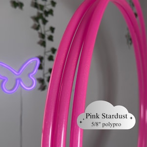Puede incluir: Aros de hula-hoop rosa brillante hechos de polipropileno de 5/8". Los aros son curvos y brillantes, con una luz con forma de mariposa de color púrpura neón en el fondo. El texto "Pink Stardust" está en un gráfico con forma de nube.