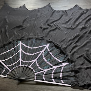 May include: A large black hand fan featuring a light pink spiderweb pattern printed on the fabric. The fan has a long, flowing black fabric veil attached to the top edge, which creates a dramatic, layered effect when the fan is fully opened.