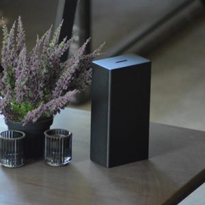 May include: A black, rectangular money box with a slot on top. The money box is made of a matte material and sits on a wooden surface. A potted plant with purple flowers and two glass candle holders are also on the table.