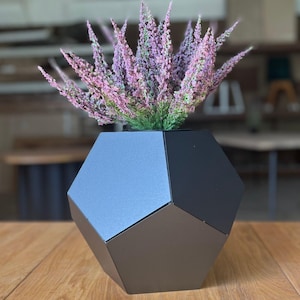 Könnte beinhalten: Eine geometrische, schwarze Metallvase mit einem vielseitigen Design, die eine lebendige Anordnung von lila und grünen Blumen enthält. Die Vase steht auf einer Holzoberfläche und schafft eine moderne, minimalistische Ästhetik. Die Blumen sorgen für einen Farbtupfer.