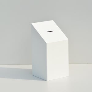 May include: A white, geometric money box with a slanted top and a narrow slot. The box is rectangular with a clean, minimalist design. The background is a plain, light gray.