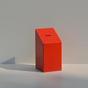 May include: A bright red, rectangular box with a slanted top and a slot. The box is a solid color and sits on a white surface. The background is a light gray.