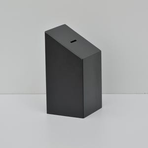 May include: A black, rectangular box with a slanted top and a narrow slot. The box is made of a solid material and stands upright on a white surface. The background is a plain, light gray.