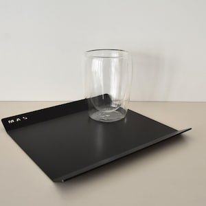 May include: A black rectangular tray with a double-walled glass tumbler. The tray has a slightly raised edge and the letters "MAS" in white. The glass is clear and has a rounded shape. The tray is on a light-coloured surface.