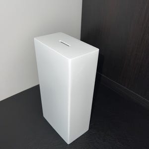 May include: A white rectangular donation box with a slot on top. The box is made of a solid material and has a smooth, matte finish. The box is positioned against a white and dark brown background.