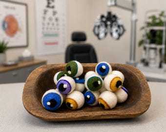 May include: A collection of round, felted wool balls designed to resemble human eyeballs, featuring white sclera and irises in various colors including blue, green, purple, and yellow, all contained within a rustic, oval-shaped wooden dough bowl.