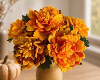 May include: A bouquet of felt flowers in shades of yellow and orange, arranged in a small, cream-colored vase. The flowers have green leaves and are set on a wooden table. A small pumpkin and a dried leaf are also visible.