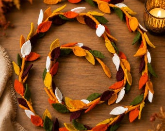 May include: A decorative garland featuring felt leaves in shades of orange, yellow, dark red, green, and white. The leaves are attached to a central orange cord, arranged in a spiral pattern to create a continuous, flexible, and textured autumn-themed strand.