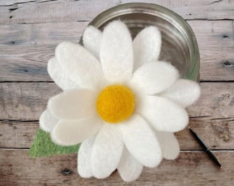 May include: A felted daisy flower with white rounded petals and a bright yellow center. The flower features a single green felt leaf attached to the base. The object is positioned next to a clear glass jar and a black pen on a wooden surface.