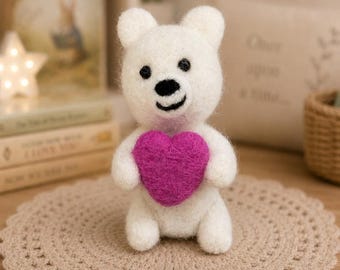 May include: A small, white, needle-felted bear figurine with black eyes and a black nose, holding a bright magenta heart made of felt. The bear is sitting upright, featuring a soft, textured wool appearance and a simple, rounded facial expression.