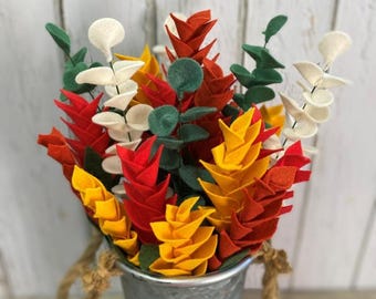 May include: A bouquet of felt flowers in a galvanized metal bucket. The flowers are in shades of red, yellow, orange, white, and green. The bucket has a rope handle. The background is a white wooden wall.