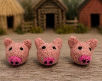 May include: Three needle-felted pig heads made from light pink wool. Each pig head features two small pointed ears, two black circular eyes, and a bright pink snout with two black nostrils. The figures are arranged in a horizontal row.