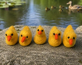 May include: Five small, yellow needle-felted ducklings arranged in a row. Each duckling features a bright orange felt beak and two small black bead eyes. The figures have a textured, fuzzy appearance and are positioned on a large, grey stone.