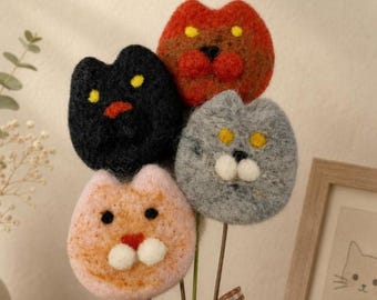 May include: A bouquet of four needle-felted cat heads in orange, black, gray, and rust colors, each featuring yellow or black eyes and white or red noses. The cat heads are attached to thin stems and tied together with a brown and white gingham ribbon.