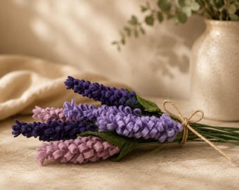 May include: A bundle of felt lavender stems in shades of purple and pink, tied with twine. The faux flowers have green leaves and are arranged on a light-colored surface. A beige vase with greenery is in the background.