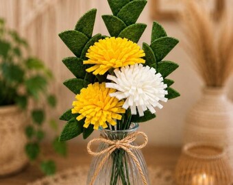 May include: A bouquet of handmade felt flowers in a clear glass vase tied with twine. The arrangement features yellow and white flowers with green leaves. The vase sits on a woven mat, with other decorative items in the background.