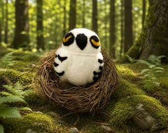 May include: A needle-felted white owl figurine with black wing markings, a black beak, and yellow-rimmed eyes. The owl is nestled inside a circular brown twig nest, featuring a soft, fuzzy texture throughout its rounded body and head.