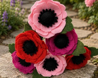 May include: A collection of felt flowers in shades of pink, red, and magenta, with black centers and green leaves. The handmade flowers are arranged on a stone surface, with a blurred garden background.