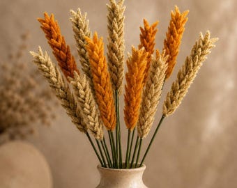 May include: A bouquet of artificial wheat stalks in a light beige ceramic vase. The stalks feature a mix of vibrant orange and natural tan colors, with textured, grain-like heads attached to thin green stems, arranged in a vertical display.
