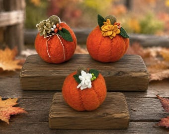 May include: Three round orange felt pumpkins with textured surfaces. Each pumpkin is topped with decorative elements including small fabric flowers, green felt leaves, and tiny berries. The pumpkins are displayed on two separate dark brown wooden blocks.