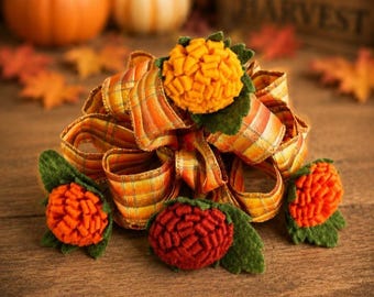 May include: A decorative bow made of orange, yellow, and brown plaid ribbon, accented with three felt flowers in shades of yellow, orange, and deep red. Each flower is surrounded by small green felt leaves, creating a textured, layered appearance.
