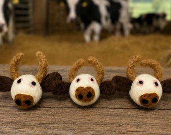 May include: Three needle-felted cow heads arranged in a row. Each cow head features a white face, dark brown ears, a brown muzzle with two black nostrils, and two curved brown horns. The texture of the felted wool is visible on each piece.