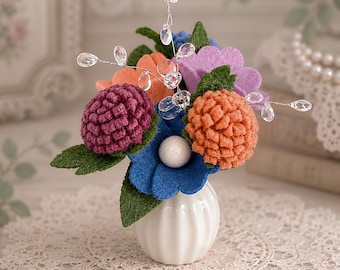 May include: A small white vase holds a colorful felt flower arrangement. The arrangement includes orange, purple, and blue flowers, with green leaves and crystal accents. The vase sits on a white lace doily.