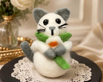 May include: A needle-felted cat figurine with white fur, grey ears, and a grey tail. The cat has black bead eyes, black whiskers, and a small grey nose. It is holding a green felted stem with a white and orange flower in its front paws.