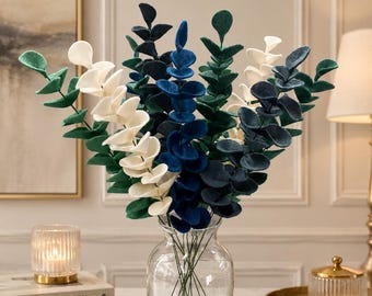 May include: A clear glass vase holds a bouquet of artificial eucalyptus. The faux eucalyptus features leaves in shades of white, green, and blue. The vase sits on a marble surface, with a candle and books in the background.