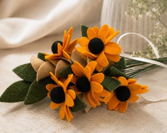 May include: A handcrafted bouquet of felt sunflowers with orange petals and black centers. The bouquet includes green leaves and tan accents, tied with a white ribbon. The flowers are arranged on a neutral surface.