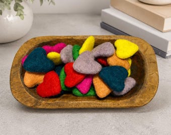 May include: A collection of small, felted wool hearts in various colors including red, yellow, orange, green, blue, pink, and grey, piled inside an elongated, oval-shaped wooden dough bowl with a dark, natural wood grain finish.