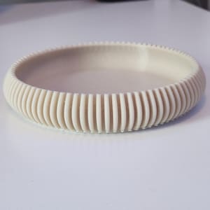May include: A beige, circular dish with a ribbed, textured exterior. The dish has a shallow interior and a rounded shape. The ribbed design creates a pattern around the edge of the dish. The dish is sitting on a white surface.