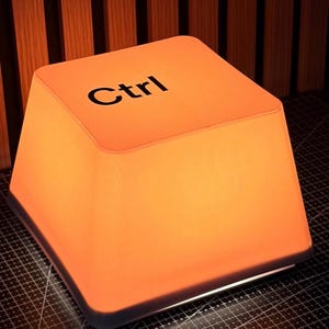 May include: A square, orange-glowing lamp shaped like a computer keyboard key. The top surface is flat with the black text 'Ctrl' printed in a sans-serif font. The lamp has a tapered, four-sided design with rounded corners and a white base.