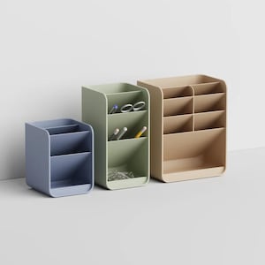 May include: Three desk organizers in blue, sage green, and beige. The blue organizer has two compartments, the green one has three compartments containing pens and scissors, and the beige one has six compartments arranged in a grid-like structure.