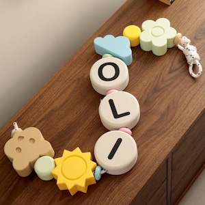 May include: A decorative garland featuring pastel-coloured silicone shapes including a light blue cloud, a yellow sun, a pale green flower, and three white circular beads printed with the black letters O, L, and I, all strung on a white rope.