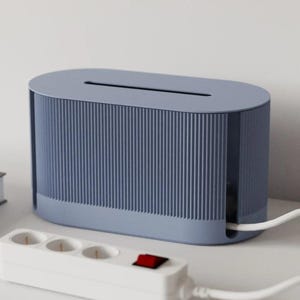 May include: A blue cable management box with a slotted top and vertical ribbed texture. A white power strip with three outlets and a red power switch is in the foreground. A white cable is plugged into the box.