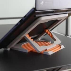 May include: A gray and orange laptop stand, designed to elevate a laptop for improved ergonomics. The stand features a foldable design with a series of orange ridges for stability and a gray frame. The laptop is black.