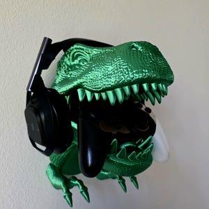 Könnte beinhalten: Eine grüne, dinosaurierförmige Wandhalterung für Gaming-Zubehör. Sie hält ein schwarzes Headset, einen schwarzen Controller und einen weißen Controller. Der Dinosaurier hat scharfe Zähne und Krallen.