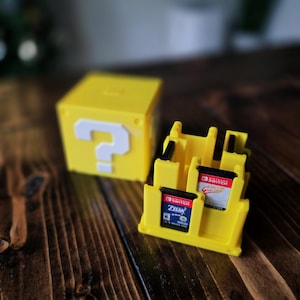 May include: A yellow question mark block and a yellow game cartridge holder. The holder contains two Nintendo Switch game cartridges. The question mark block is a cube with a white question mark on one side. The items are on a dark wooden surface.