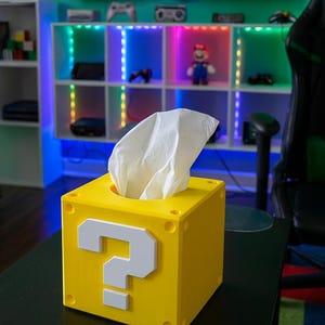 May include: A yellow tissue box, designed as a question mark block from the Super Mario video game series. The box features a white question mark and white tissues emerging from the top. It is placed on a black table, with a shelf unit in the background.