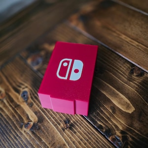 Könnte beinhalten: Ein leuchtend rosa rechteckiges Gehäuse, wahrscheinlich für eine Spielekonsole, mit einem weißen Logo, liegt auf einer dunklen Holzoberfläche. Das Logo ist ein weißes, stilisiertes Design. Das Gehäuse ist einfarbig und scheint aus Kunststoff zu sein.